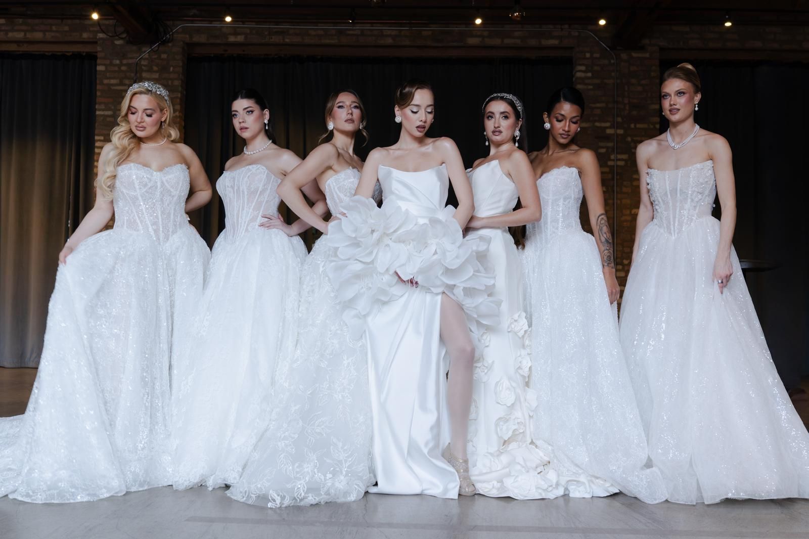 Seven women in white formal gowns posing together on a stage
