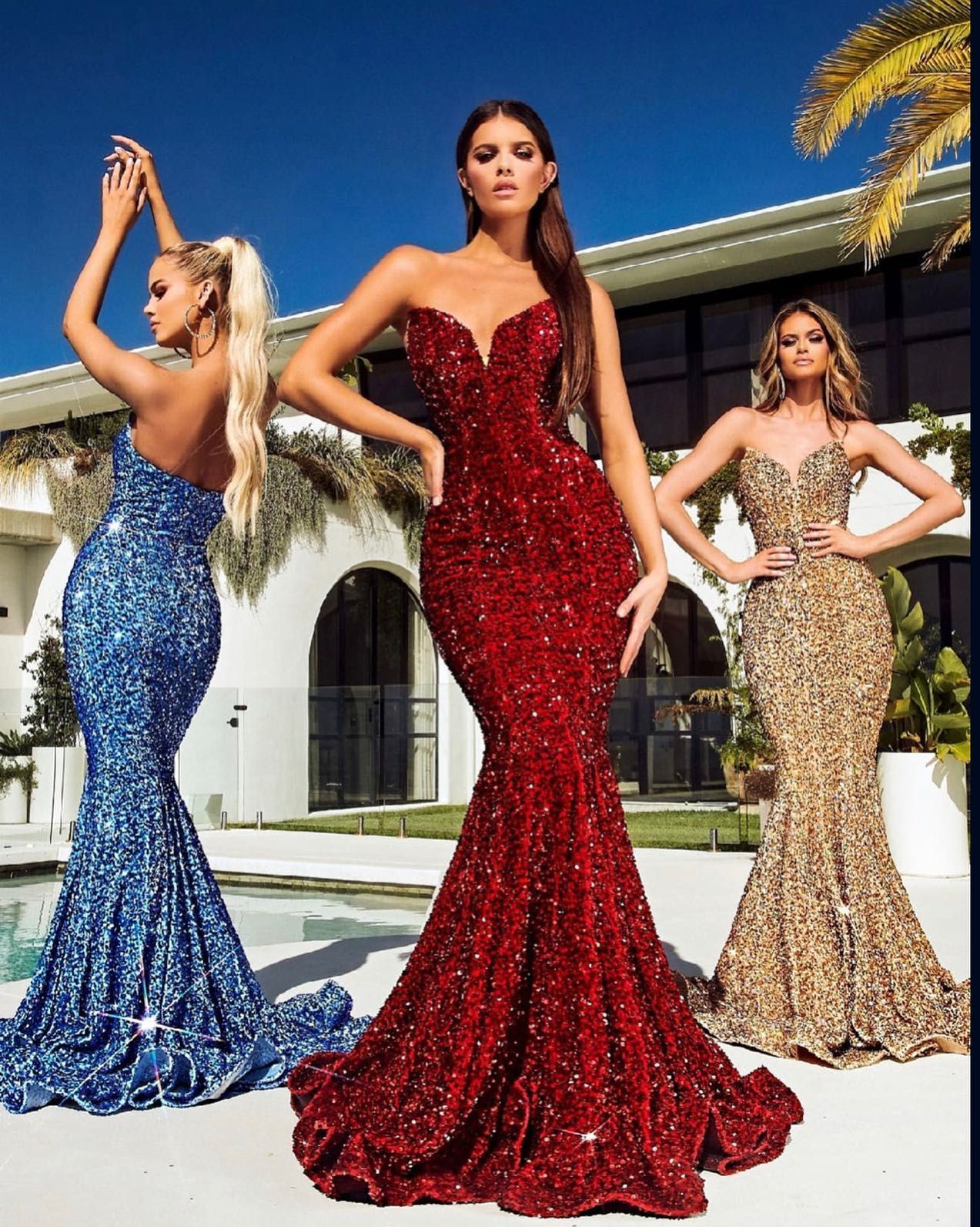 Three models pose in sparkling blue, red, and gold mermaid gowns outdoors by a villa.