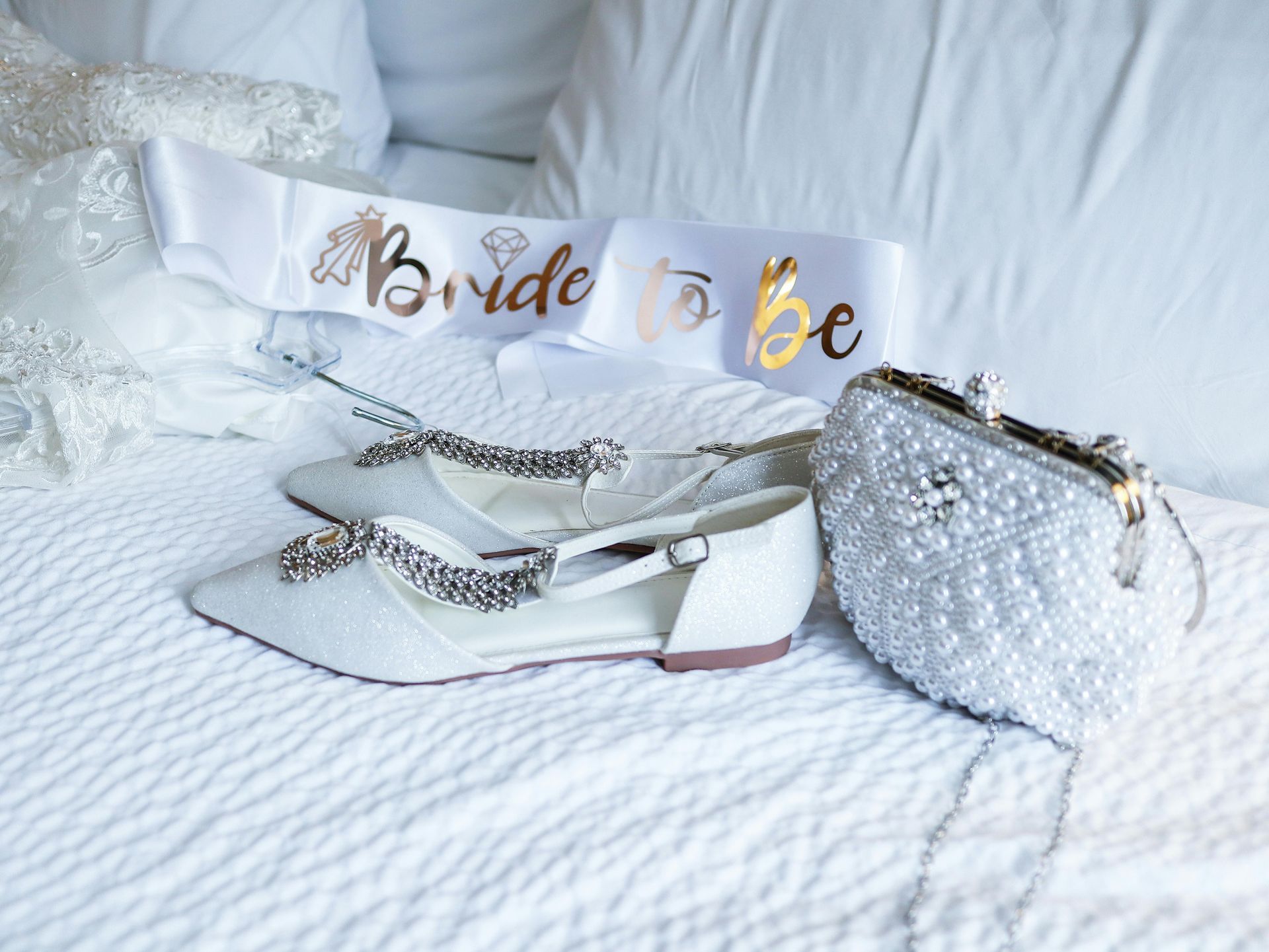 Bride gift set on white bed: pearl purse, sandals, and “Bride to Be” sash.