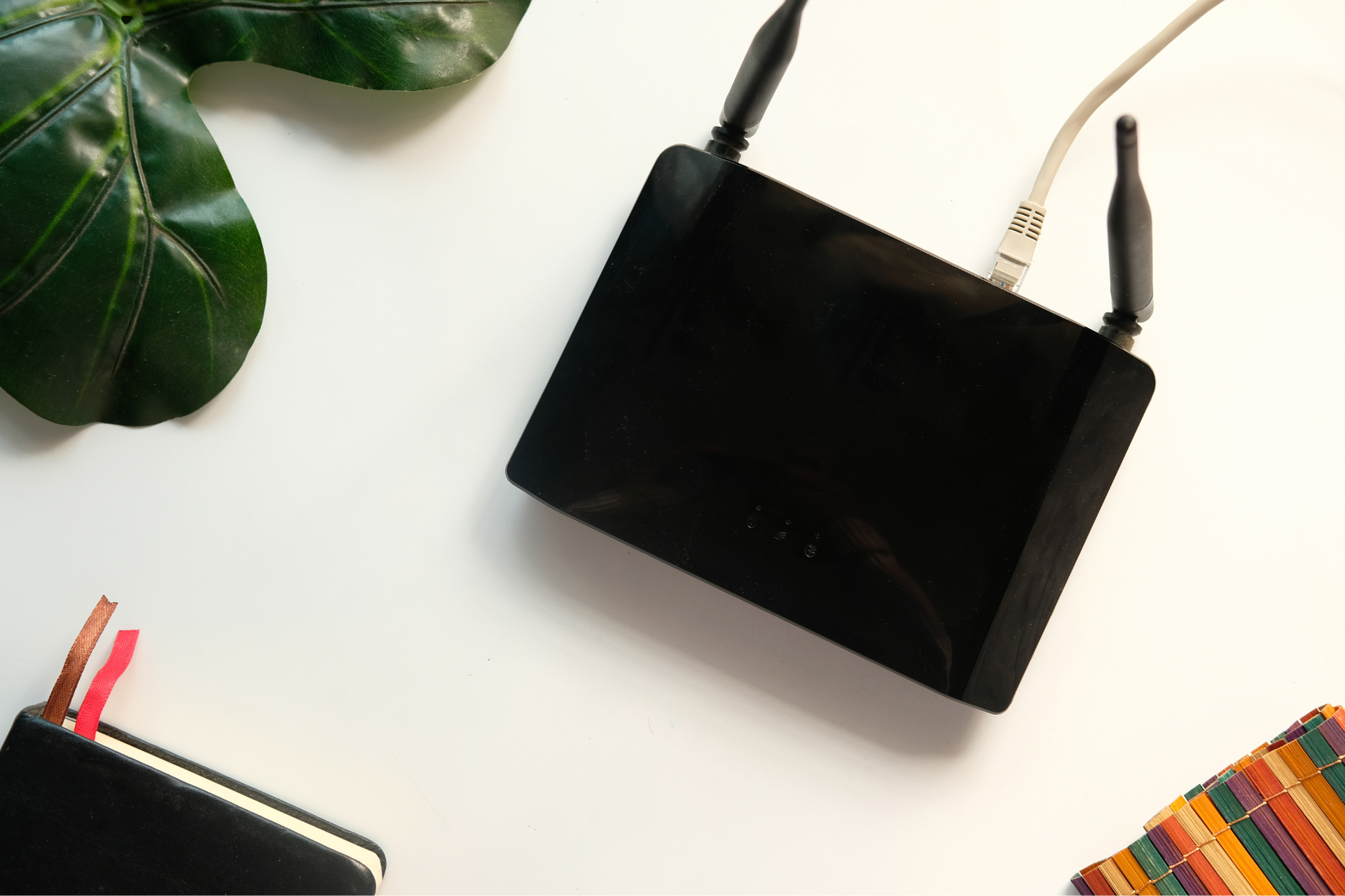 A black router is sitting on a white table next to a notebook and a plant.