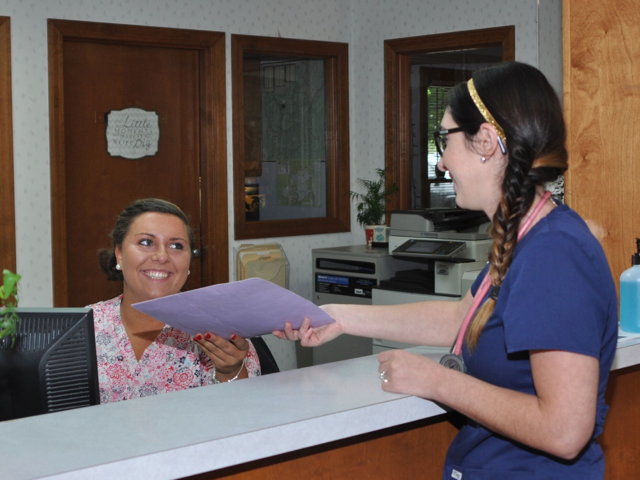 Two women at a medical desk exchanging documents and smiling at each other.