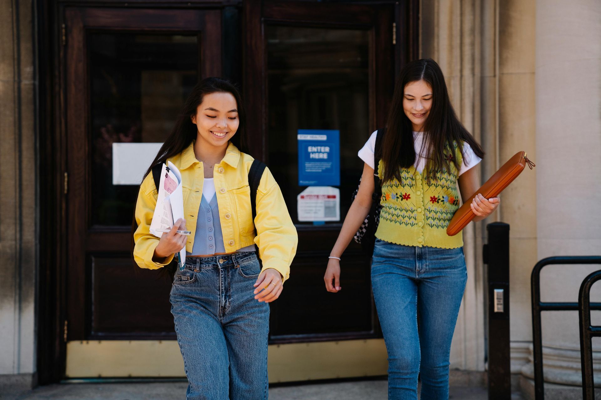 Two teenaged students are leaving a school. They are walking and smiling.