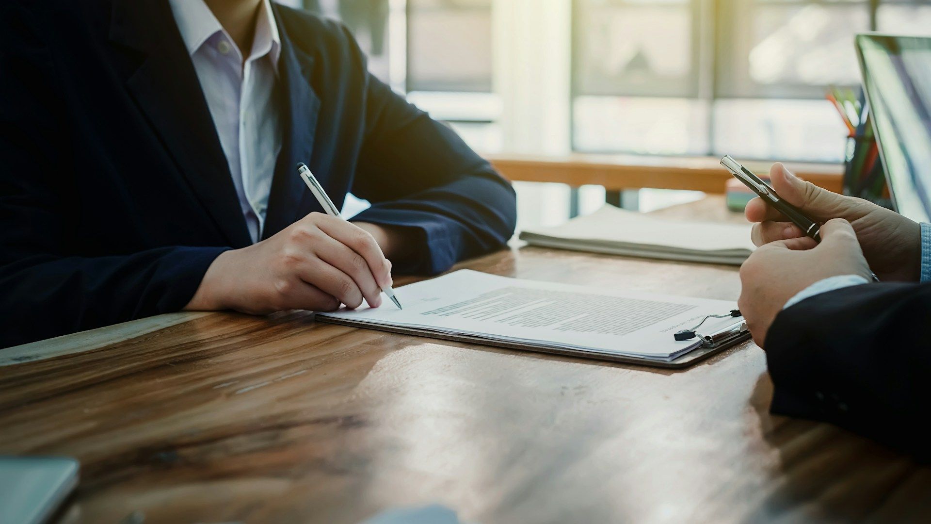 Two people in business attire discussing documents at a wooden conference table in a bright office.