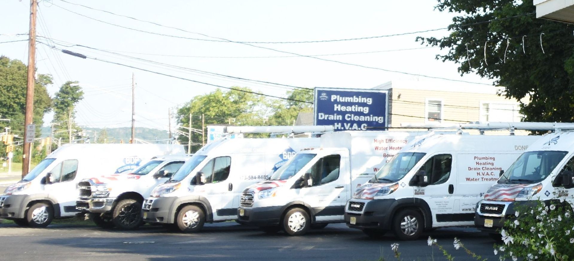 A row of white service vans parked in front of a building with a sign for plumbing, heating, and HVAC services.