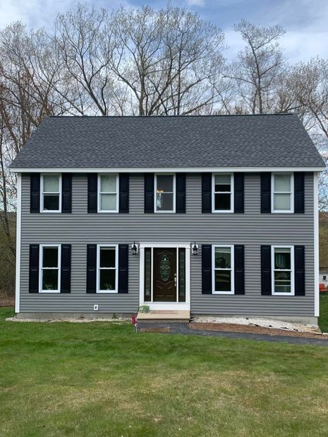 A large gray house with black shutters is sitting on top of a lush green field.