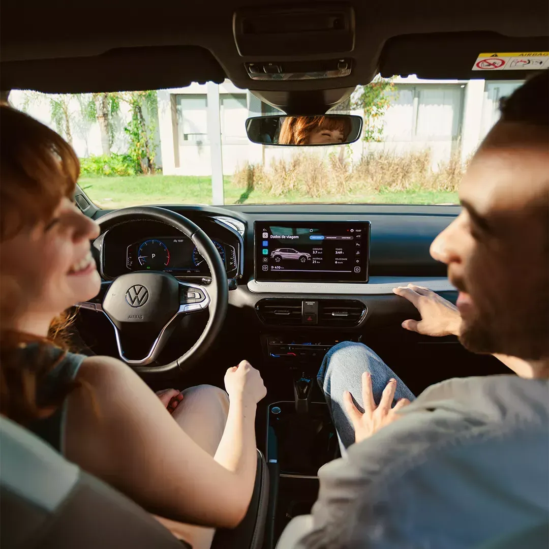Duas pessoas em um carro, olhando para o painel. Foto do interior com uma mulher sorrindo e um homem conversando.