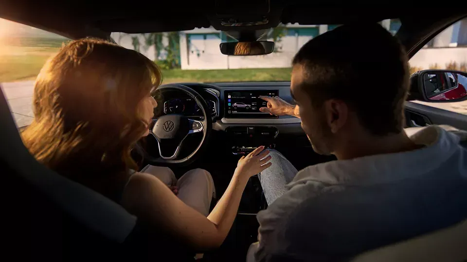 Casal no carro, ajustando a tela do sistema de informação e entretenimento. Ambiente ensolarado, lado do motorista visível.