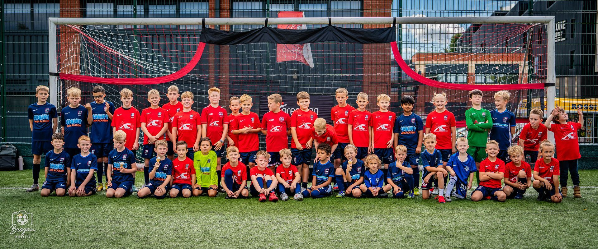 Youth football teams posing in two rows on a field in front of a goal, wearing red and blue uniforms.