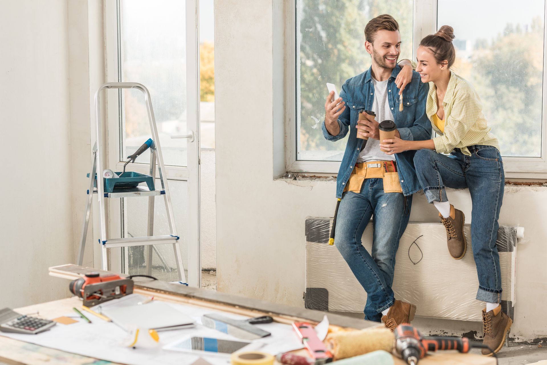 Two people take a break with drinks amid renovation tools by a window.