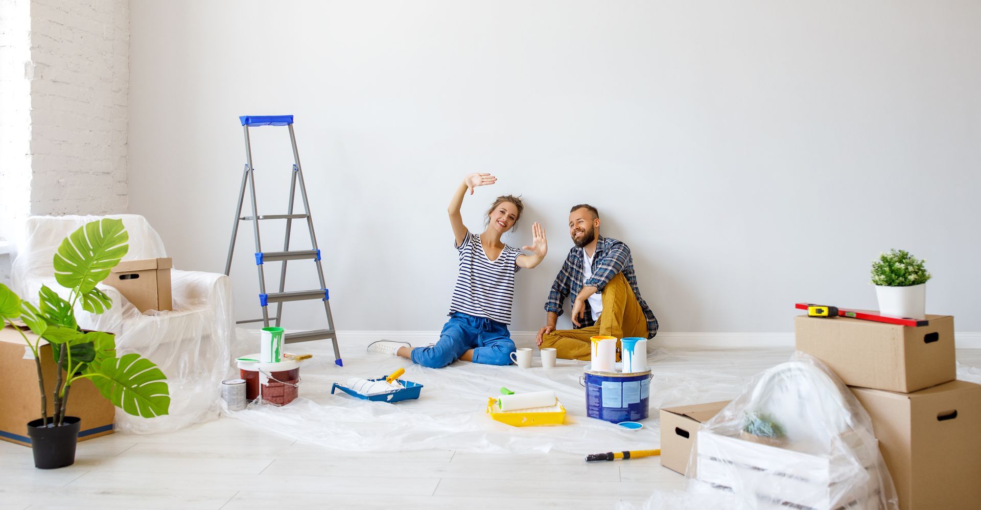 Two people sit on floor with paint supplies, ladder, and boxes in a bright room. Two people sit on floor with paint supplies, ladder, and boxes in a bright room.