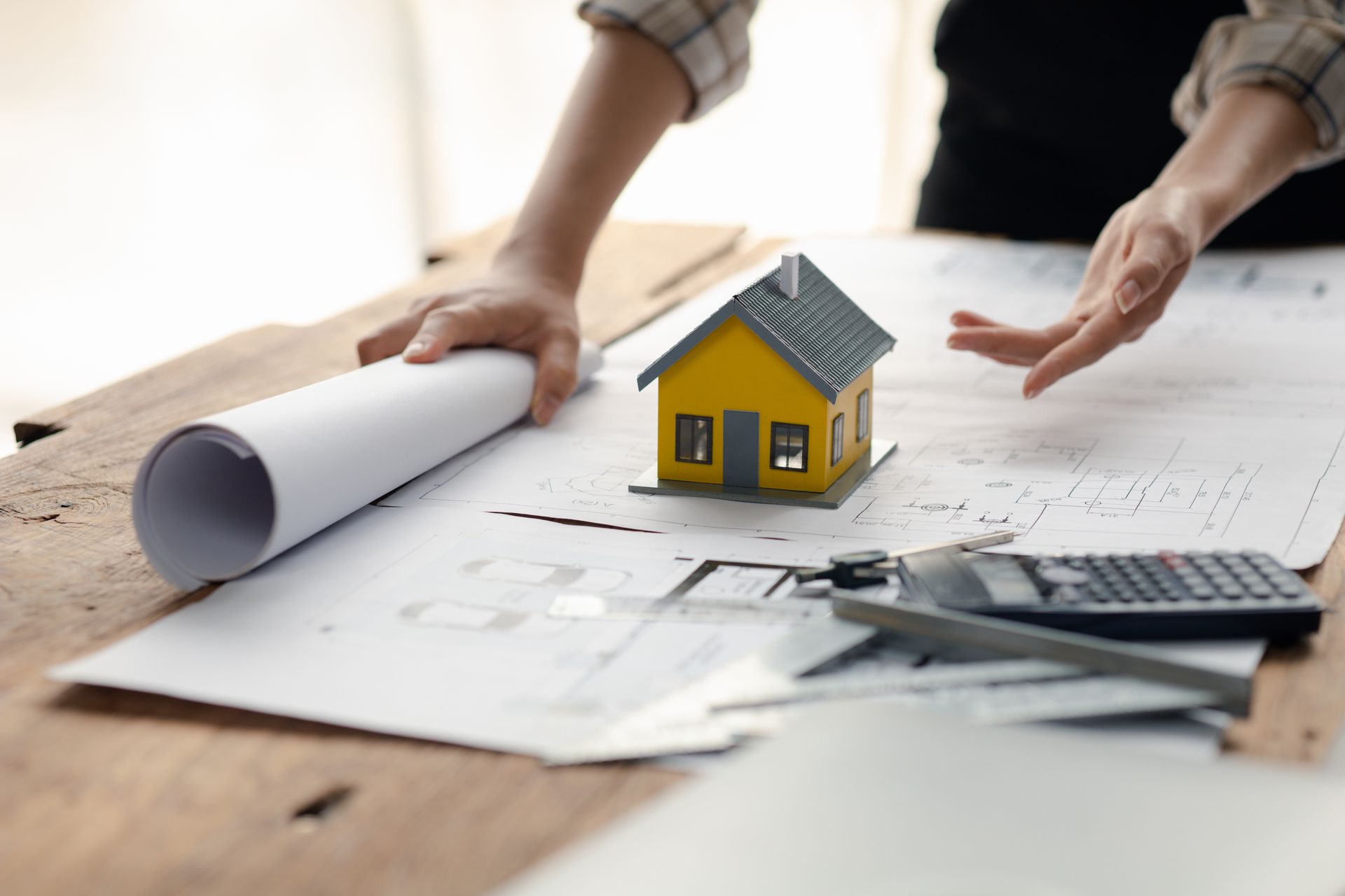 Hands over architectural plans beside a small house model and calculator on a desk