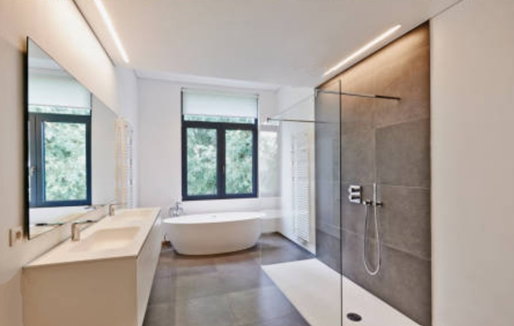 Modern bathroom with gray tiled shower, white bathtub by window, double sink, and ambient lighting.