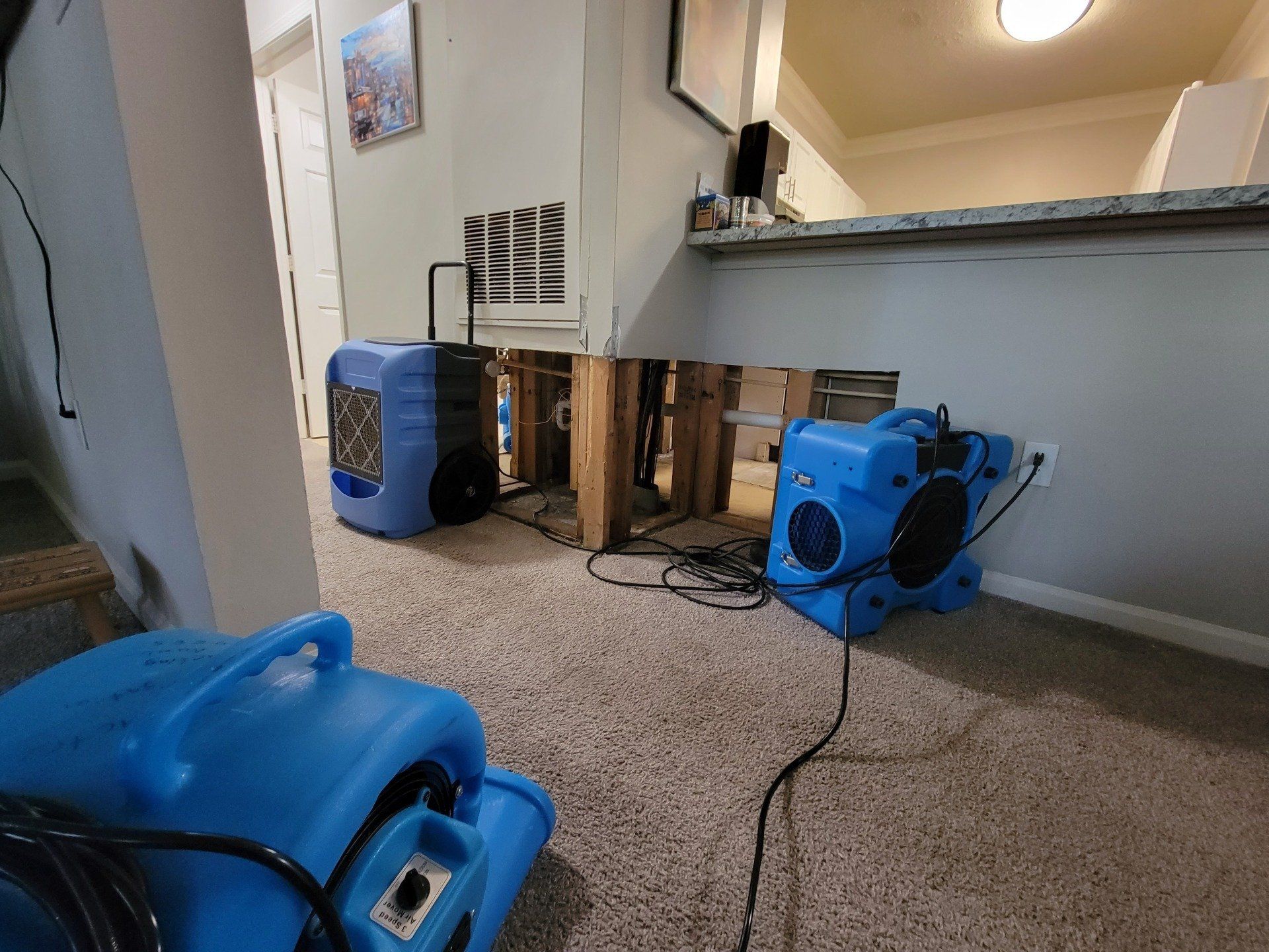 Two blue floor fans drying a damaged interior wall with exposed studs and debris under a kitchen counter