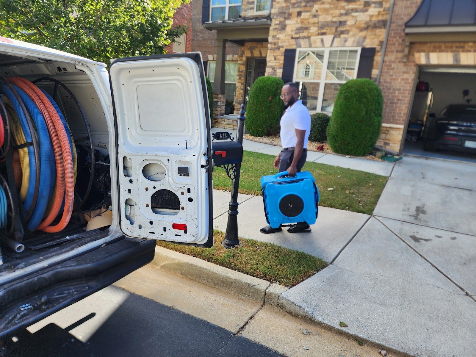 Worker carrying a blue blower on a driveway beside a truck loaded with coiled hoses, outside a home