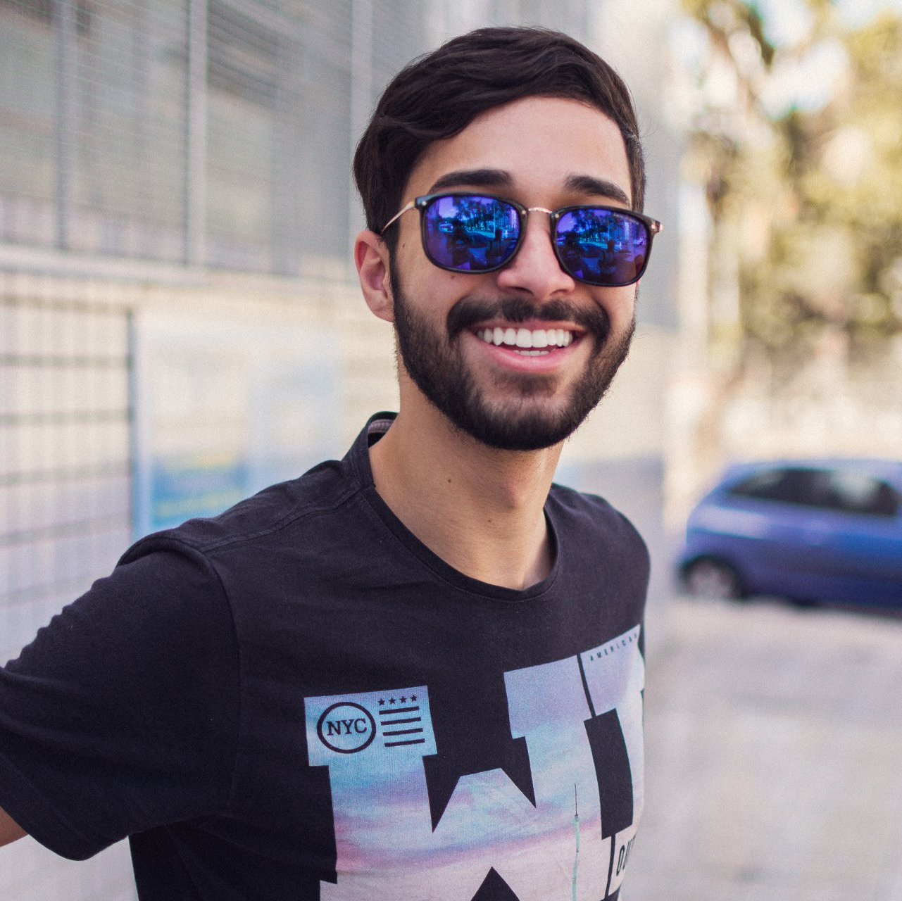 Smiling man wearing blue sunglasses and a black T-shirt, standing outdoors on a sunny street.