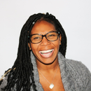 Smiling person with glasses and long braids wearing a gray sweater on a white background