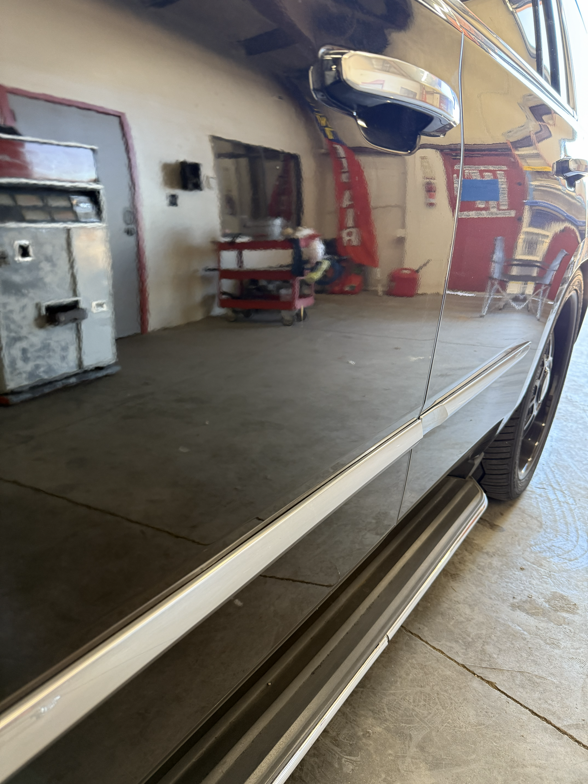 Shiny black car's side, reflecting a garage interior with equipment and a running board.