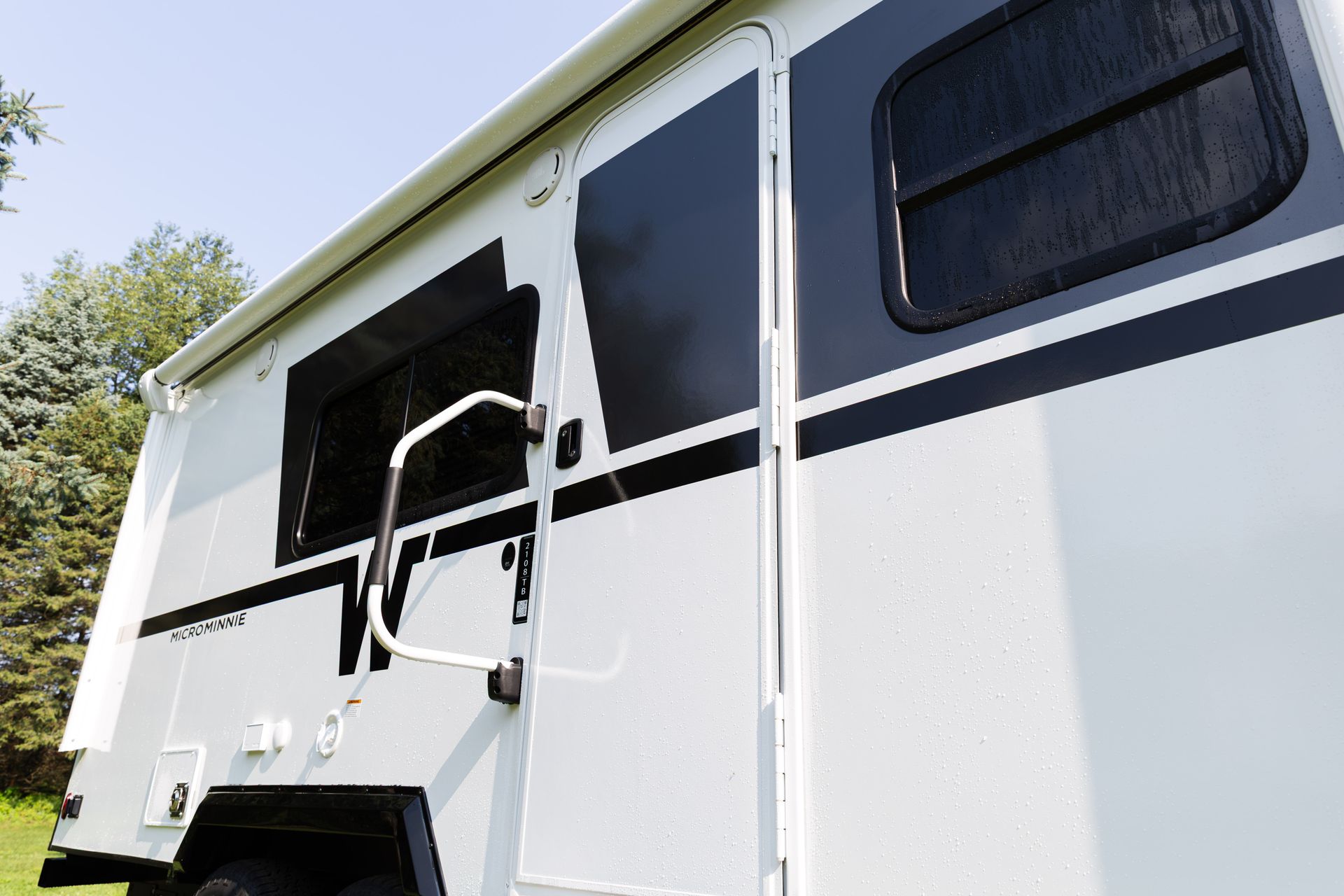 White RV side with open door, black window trim, and a metal ladder under a blue sky