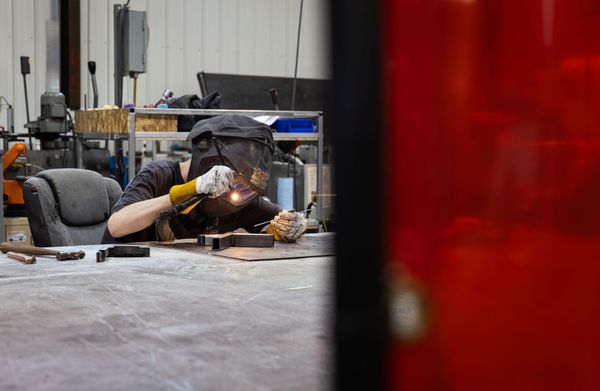 Person welding at a workbench in a workshop, with bright sparks and a red blurred foreground panel