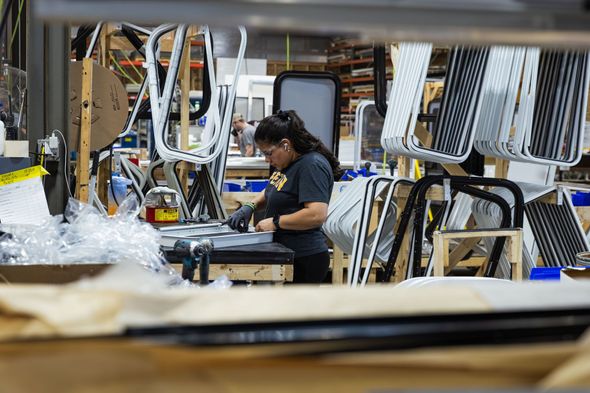 Worker assembling components on a factory line amid stacks of cables and equipment.
