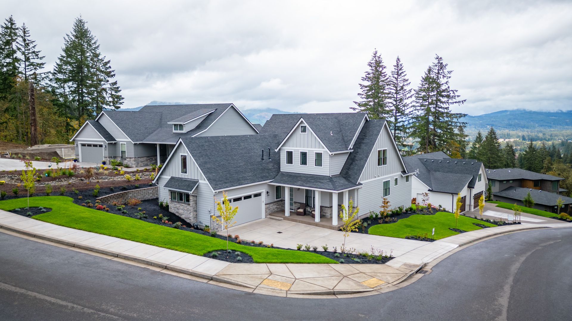 Large gray house with white trim on a corner lot, surrounded by trees and mountains under a cloudy sky