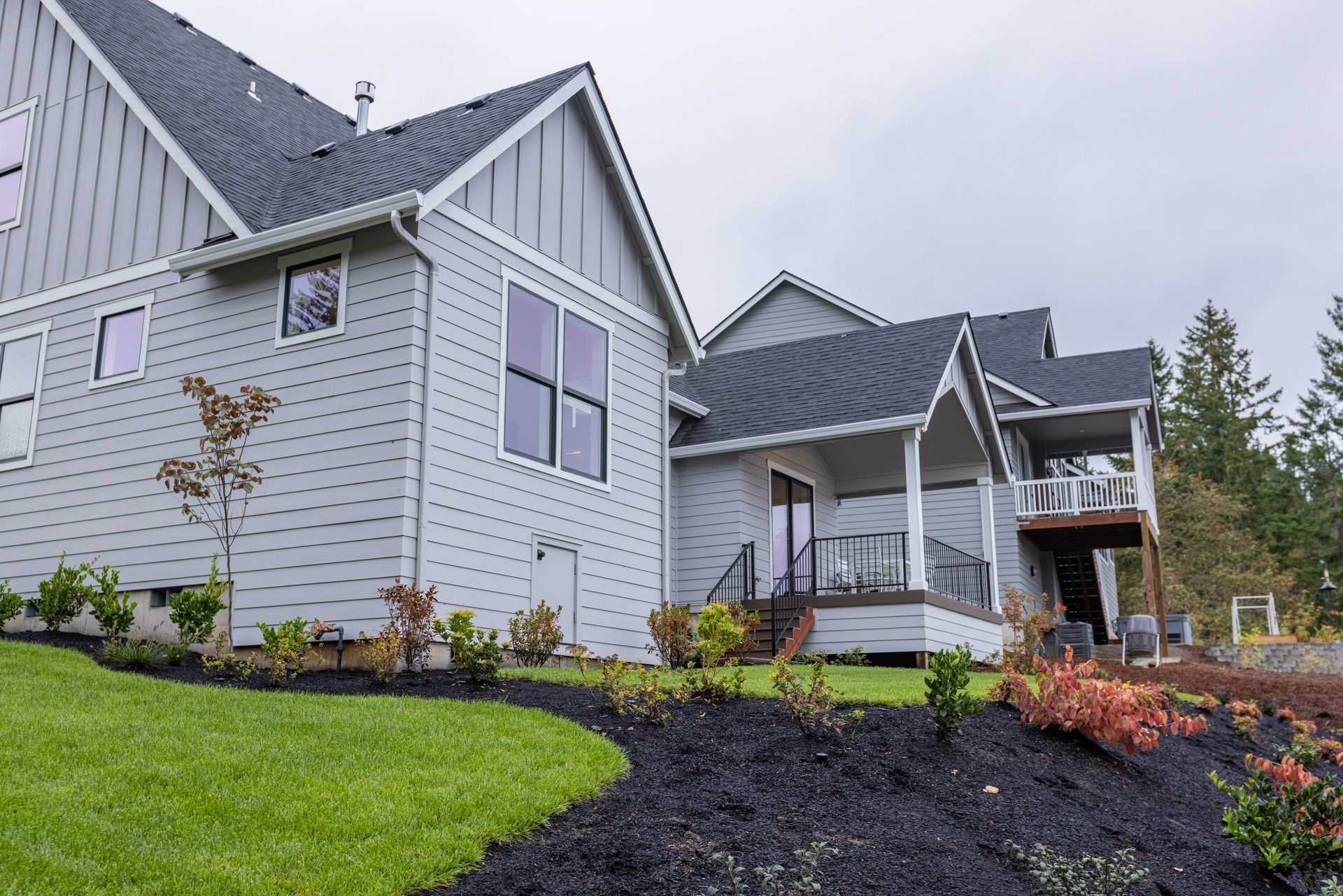 New gray suburban house with black roof, front porch, and landscaped lawn under an overcast sky