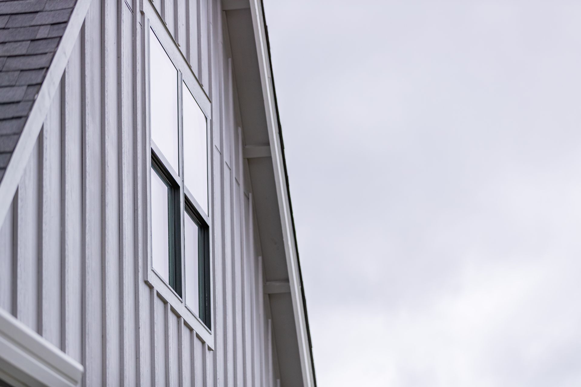 Side view of a gray house with vertical siding and a window against a cloudy sky
