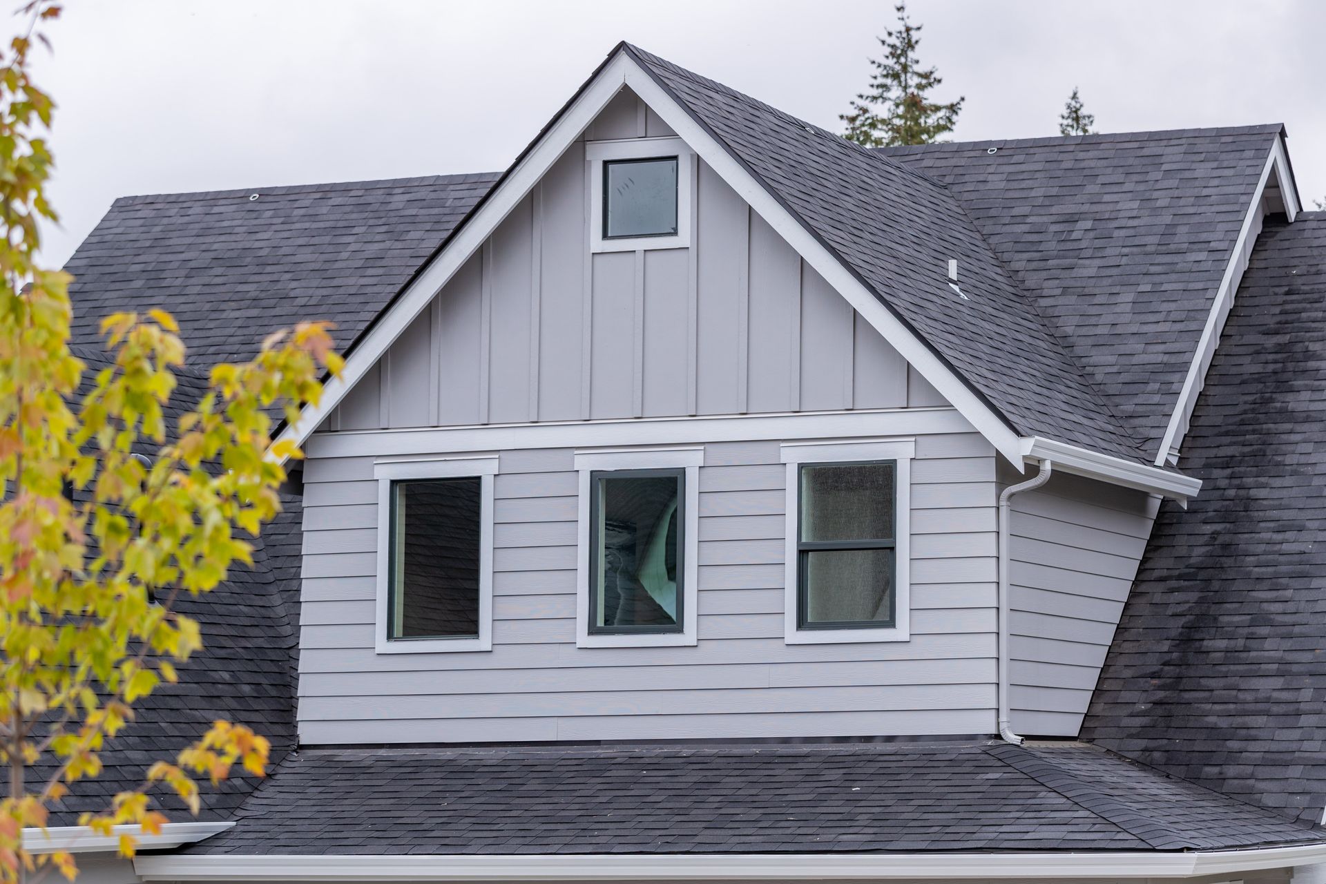 Gray house exterior with dark shingle roof and three front windows, framed by yellow leaves and pine trees