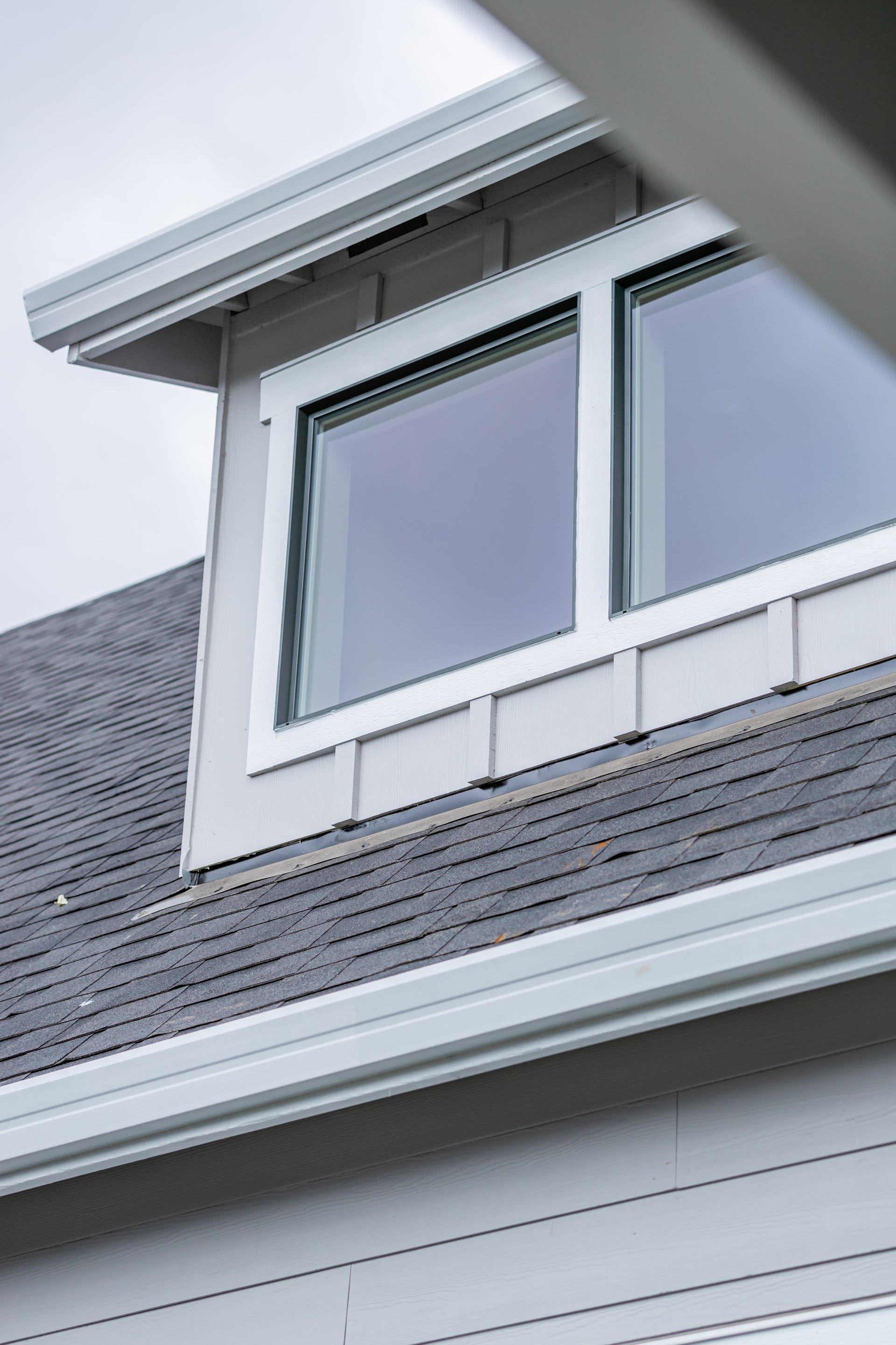 Dormer window with white trim on a gray shingled roof under an overcast sky