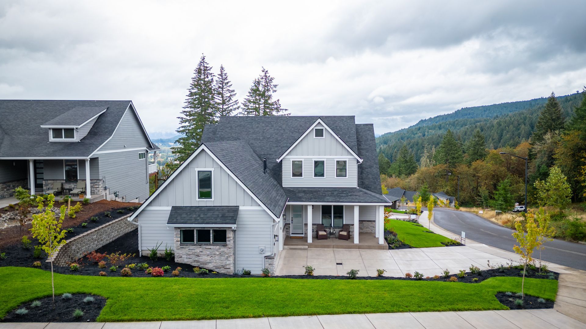 Two gray suburban houses with a green lawn, trees, and mountain backdrop under cloudy skies