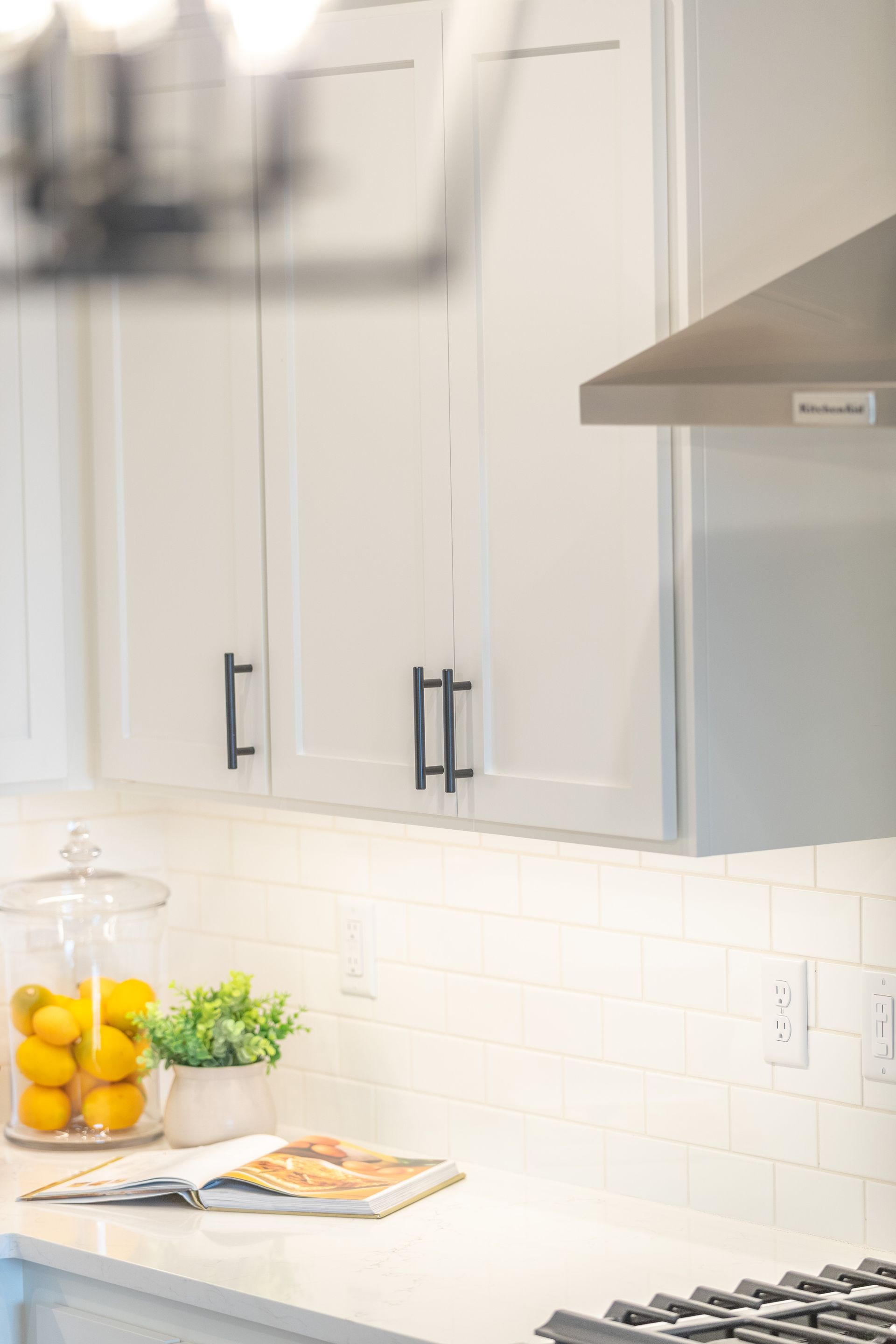 Bright white kitchen with cabinets, countertop, lemons in a glass jar, and a cookbook near a stove.