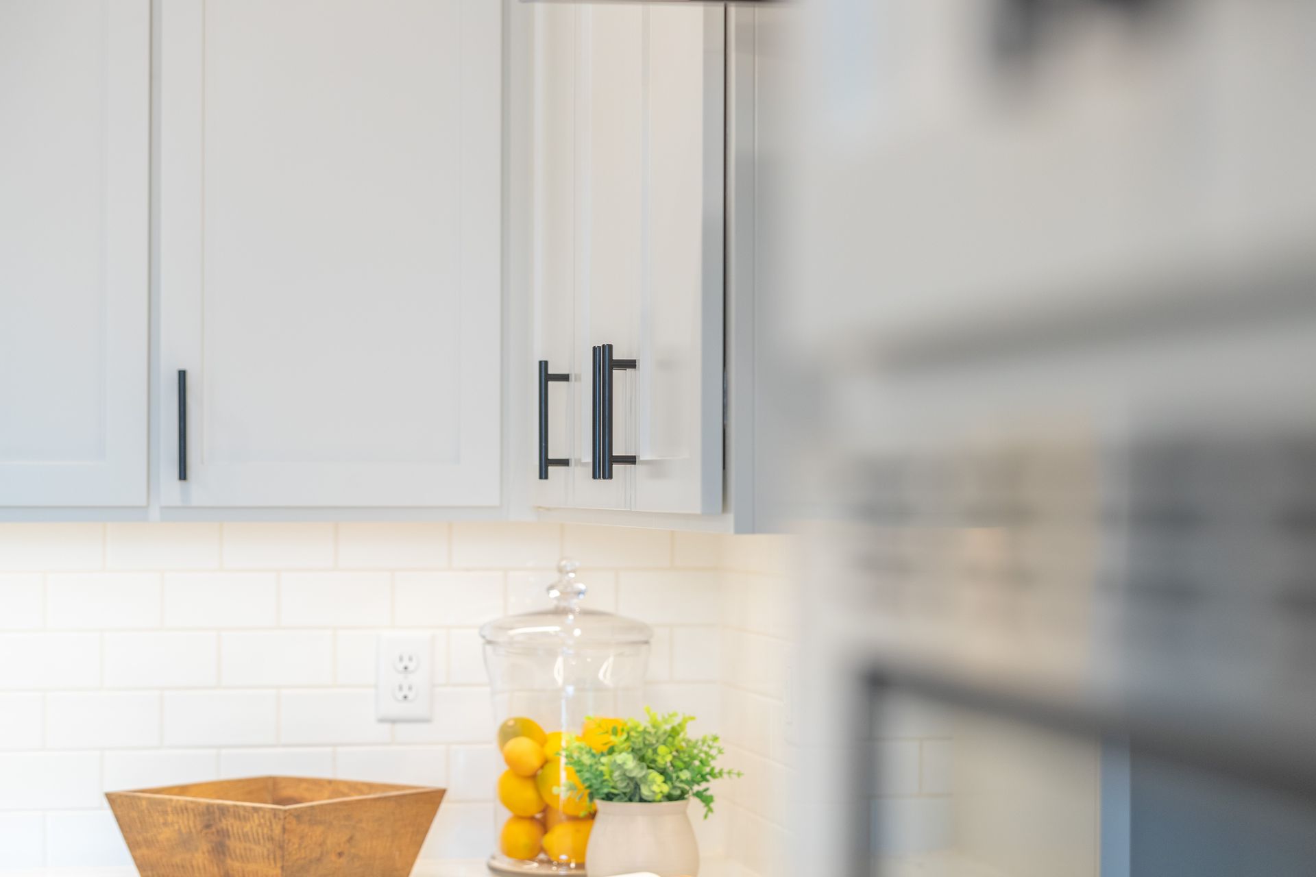 Bright kitchen counter with gray cabinets, white tile backsplash, woven basket, and glass jar of lemons and flowers