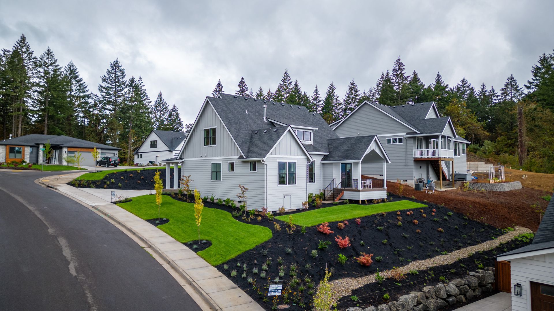Curved suburban street with white houses, manicured lawns, and a cloudy sky amid tall trees