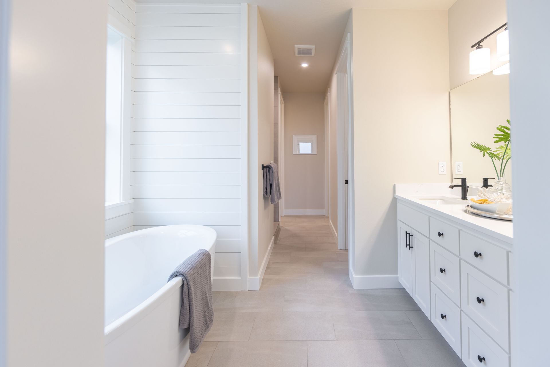 Bright modern bathroom with white vanity, tub, and a long hallway leading to a small window.