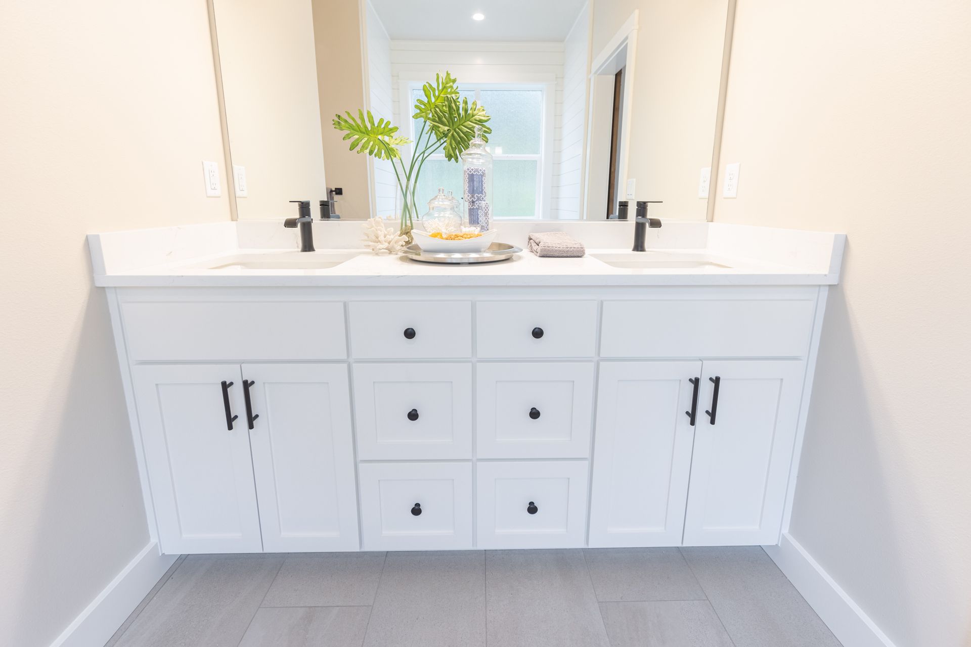 White bathroom vanity with double sinks, black fixtures, drawers, cabinet doors, and a vase of flowers.