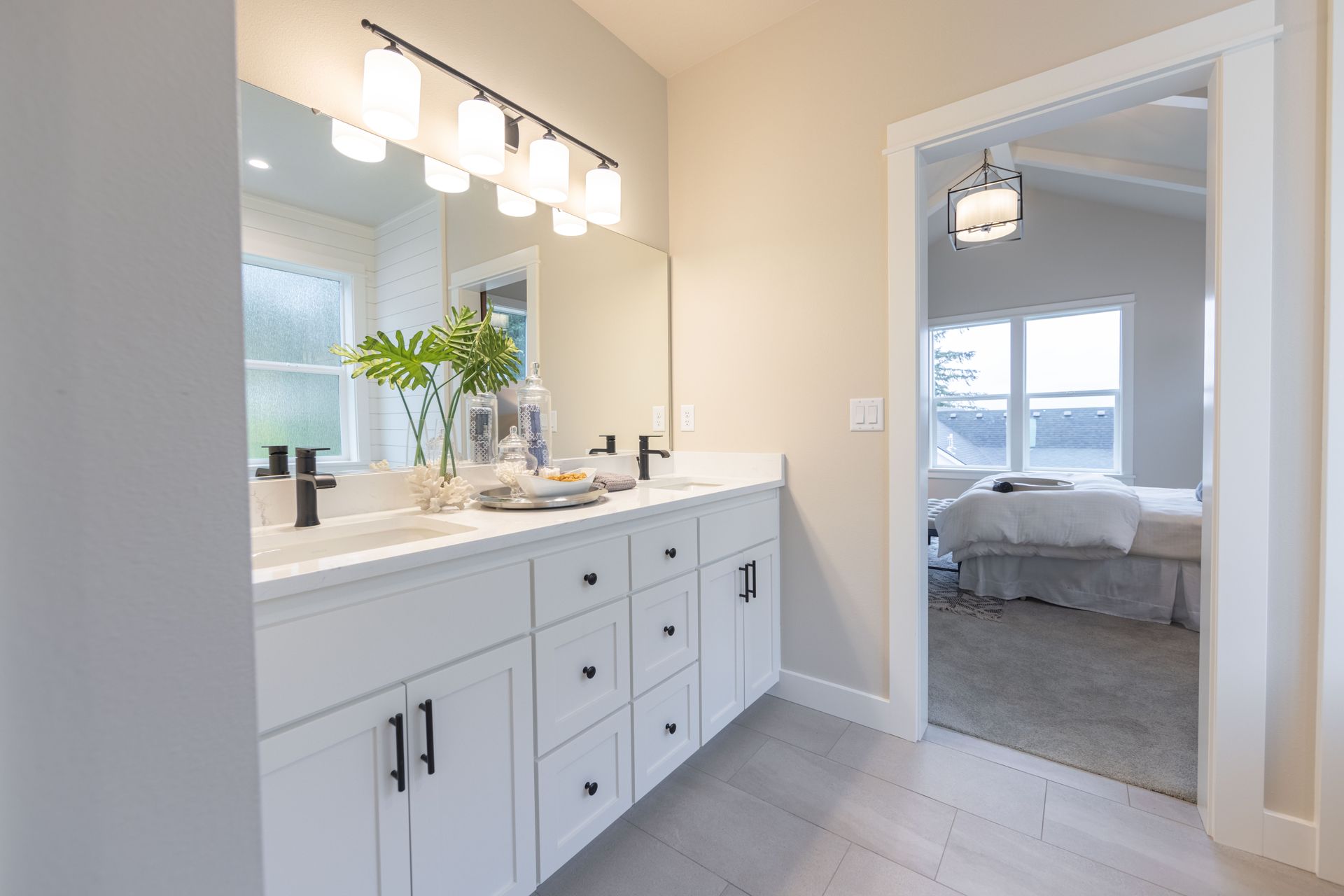 Bright bathroom with white vanity, double sinks, and mirror, opening to a bedroom with gray bedding.