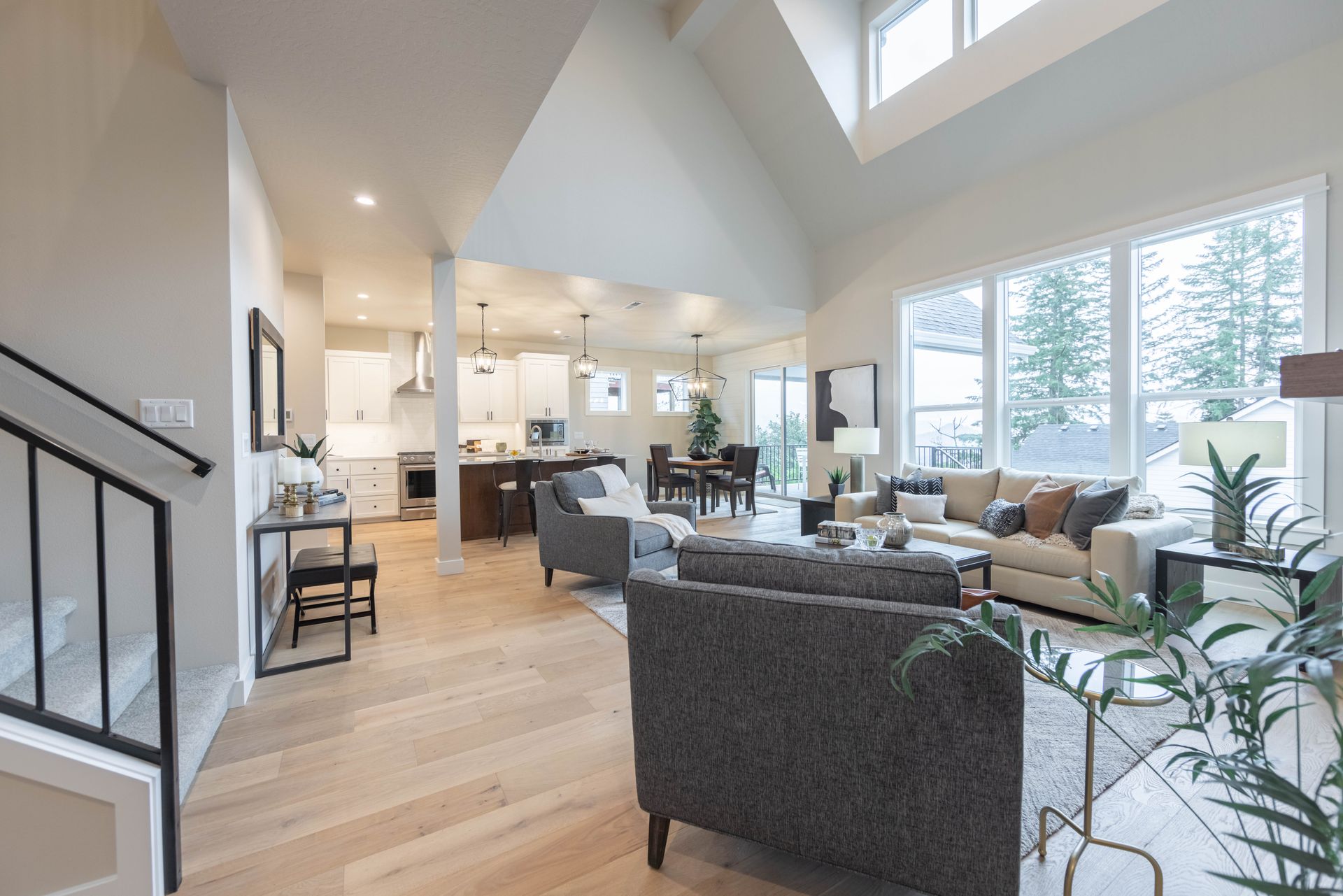 Bright open-concept living room with gray sofas, hardwood floors, and a kitchen in the background
