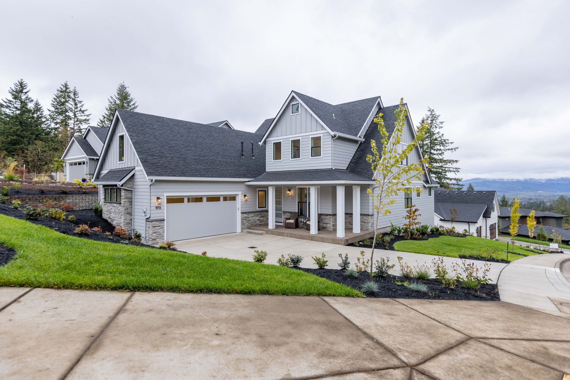 Large gray house with a three-car garage on a landscaped hillside overlooking water