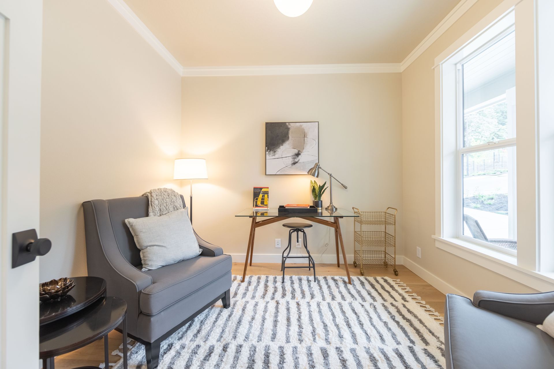 Bright home office with a gray chair, desk lamp, abstract wall art, and a striped rug by a window.