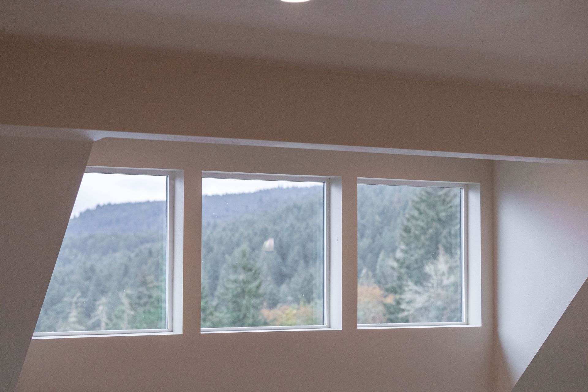 Three-pane window with a forested mountain view and slanted white walls inside a room