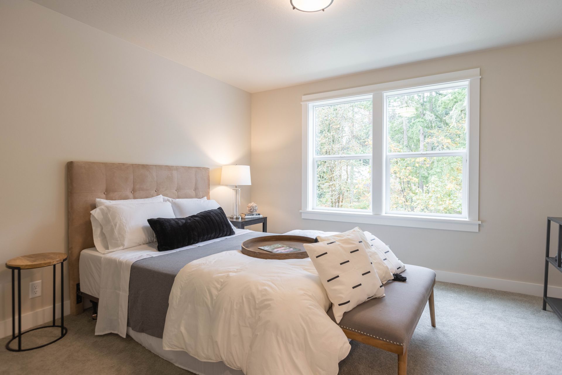 Bright bedroom with a made bed, bench, bedside lamp, and two windows with white trim