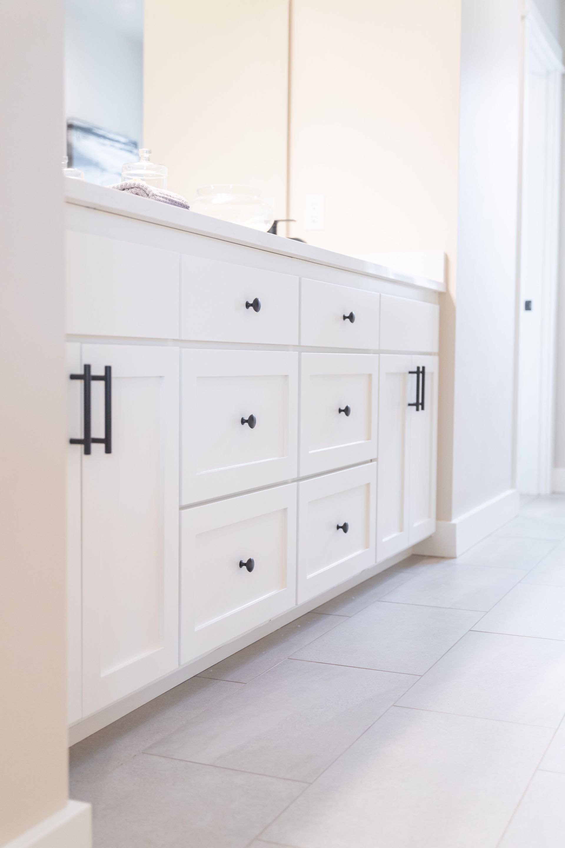 White bathroom vanity with drawers, black handles, and mirror above on a light tiled floor