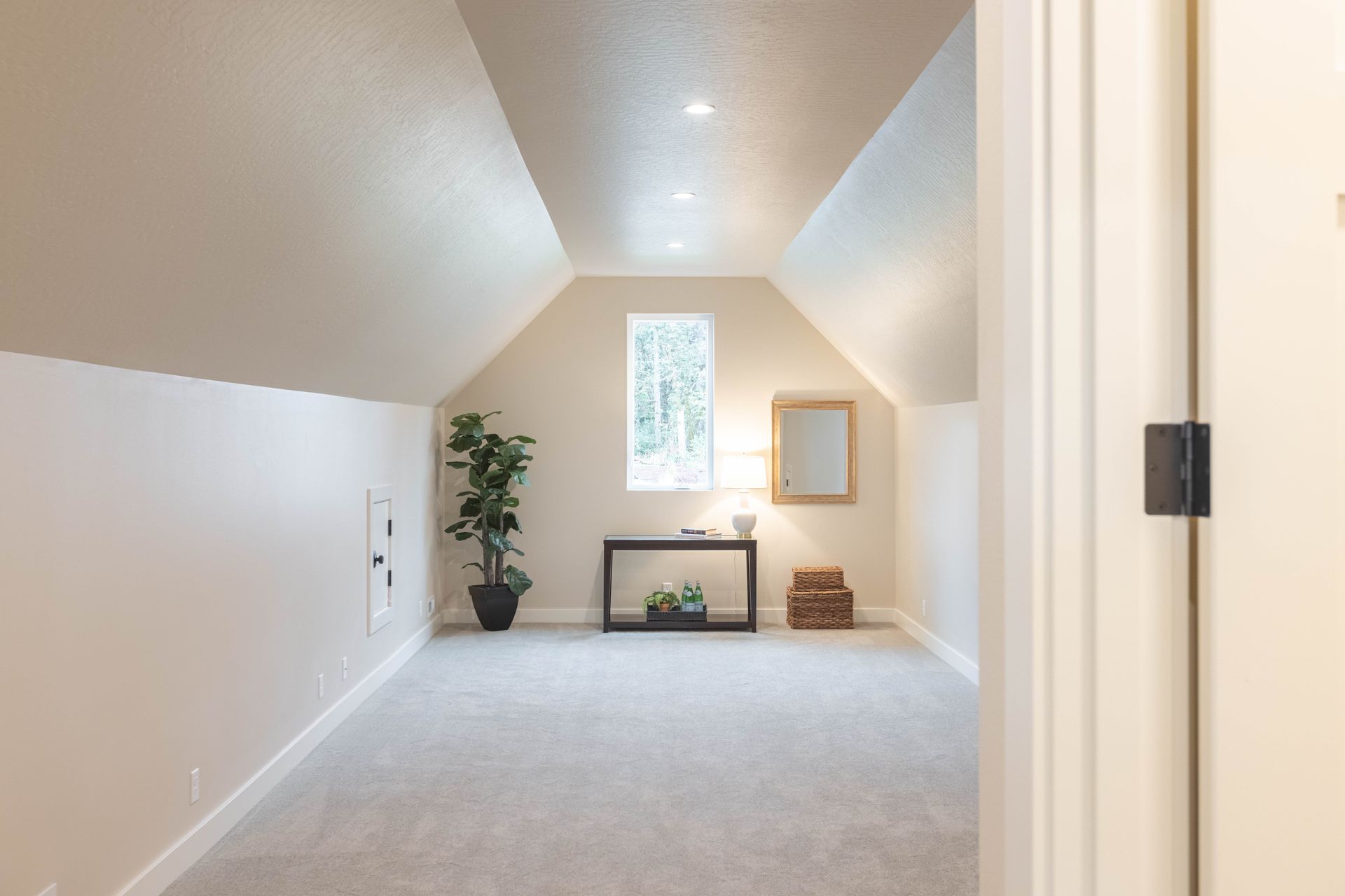 Bright carpeted attic room with sloped white ceiling, window, plants, and fireplace