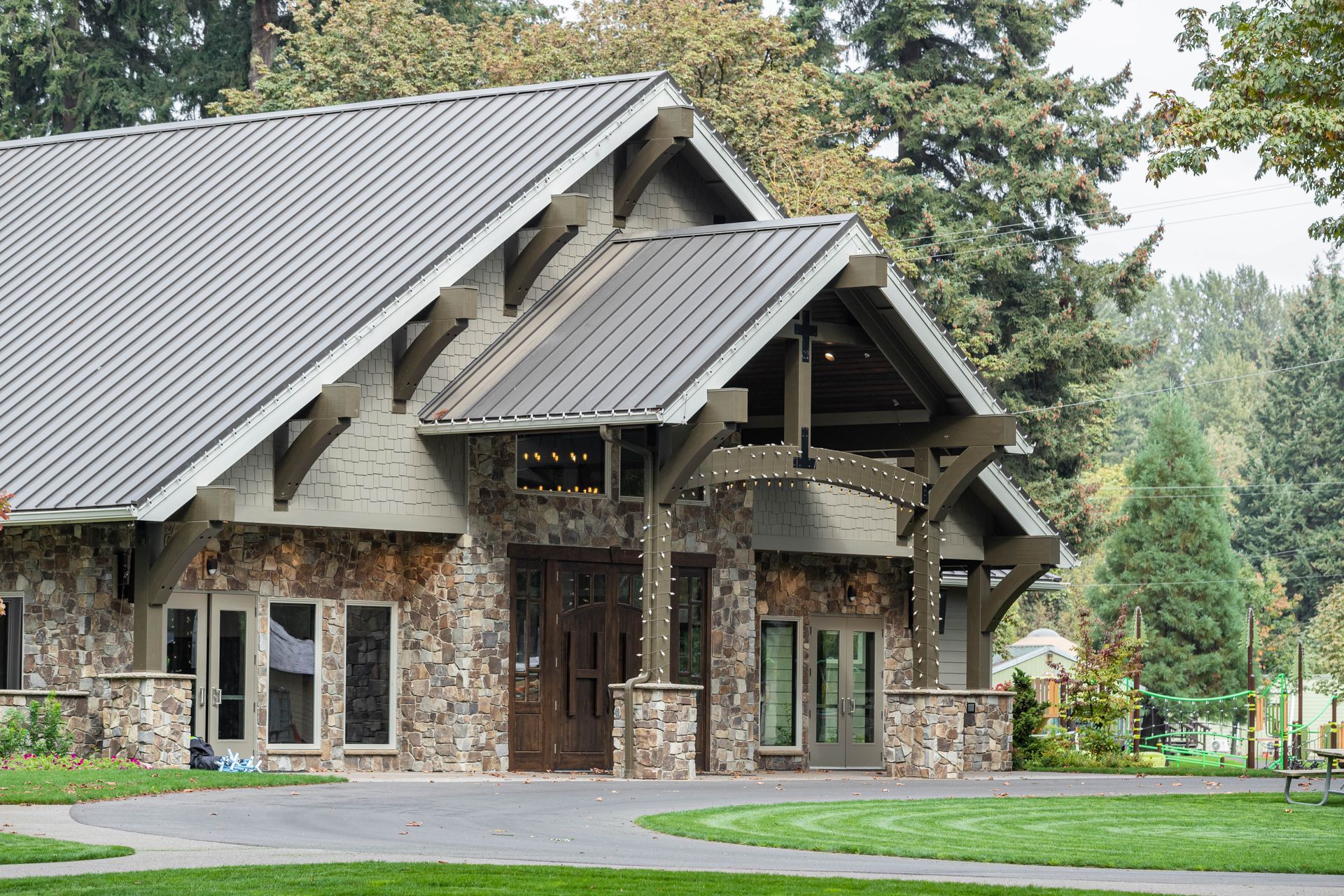 Stone lodge with gray metal roof and large windows, set among trees and a green lawn.