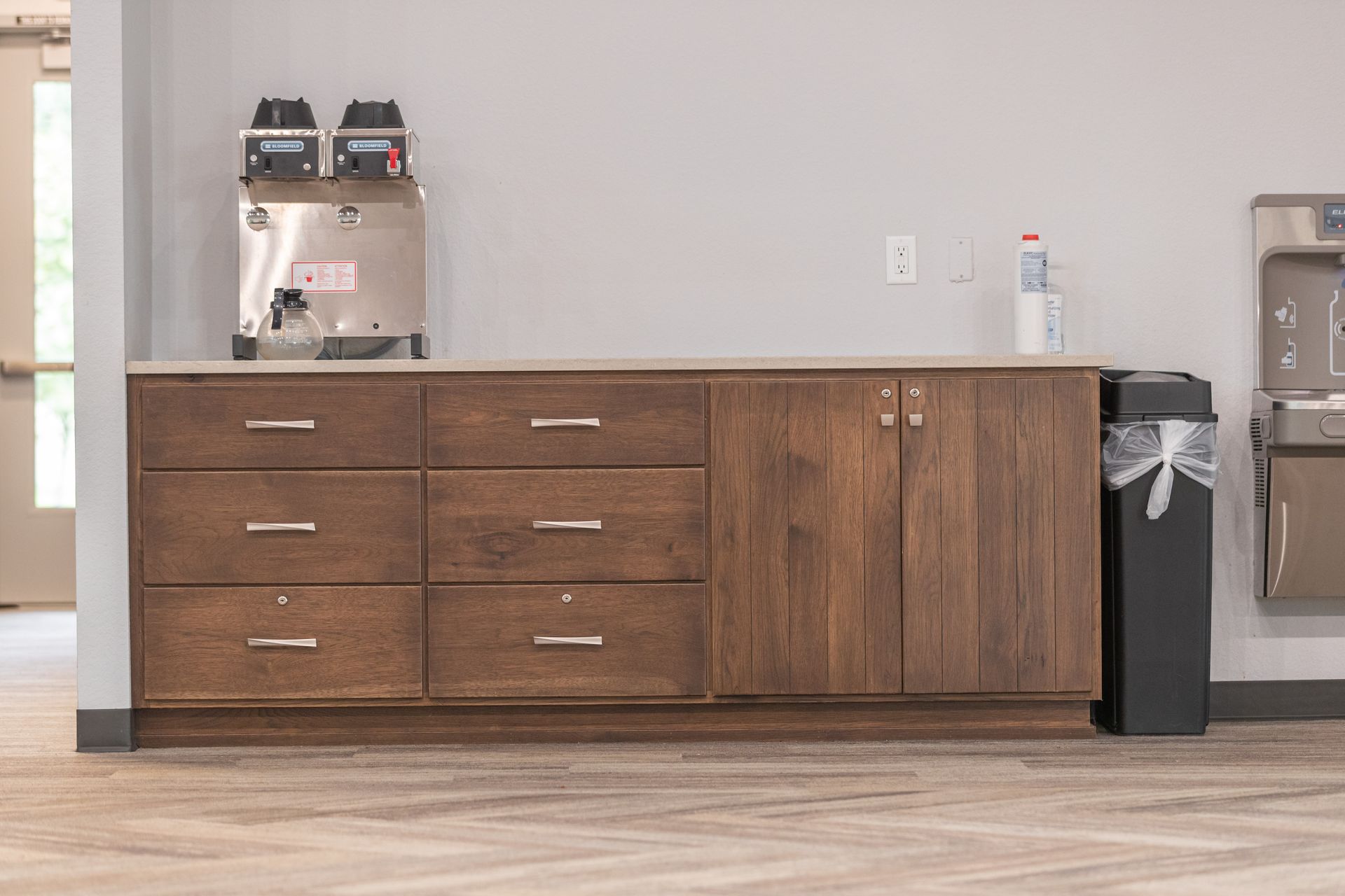 Office break room counter with drawers, coffee machine, water bottle, and trash bin against a plain wall