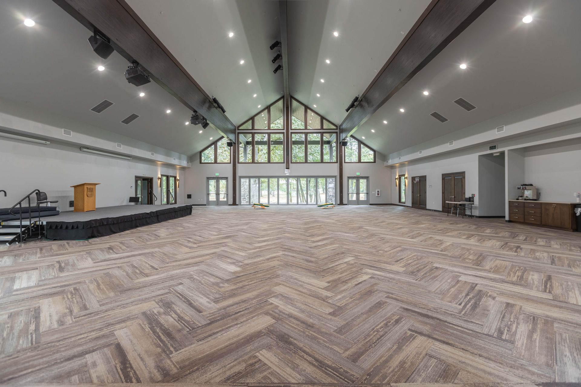 Spacious empty hall with herringbone wood floor, vaulted ceiling, and large windows at the far end