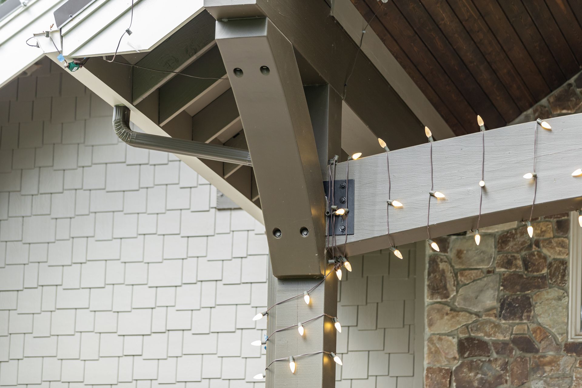 Outdoor patio roof with brown beams, string lights, and white shingle wall against stone siding