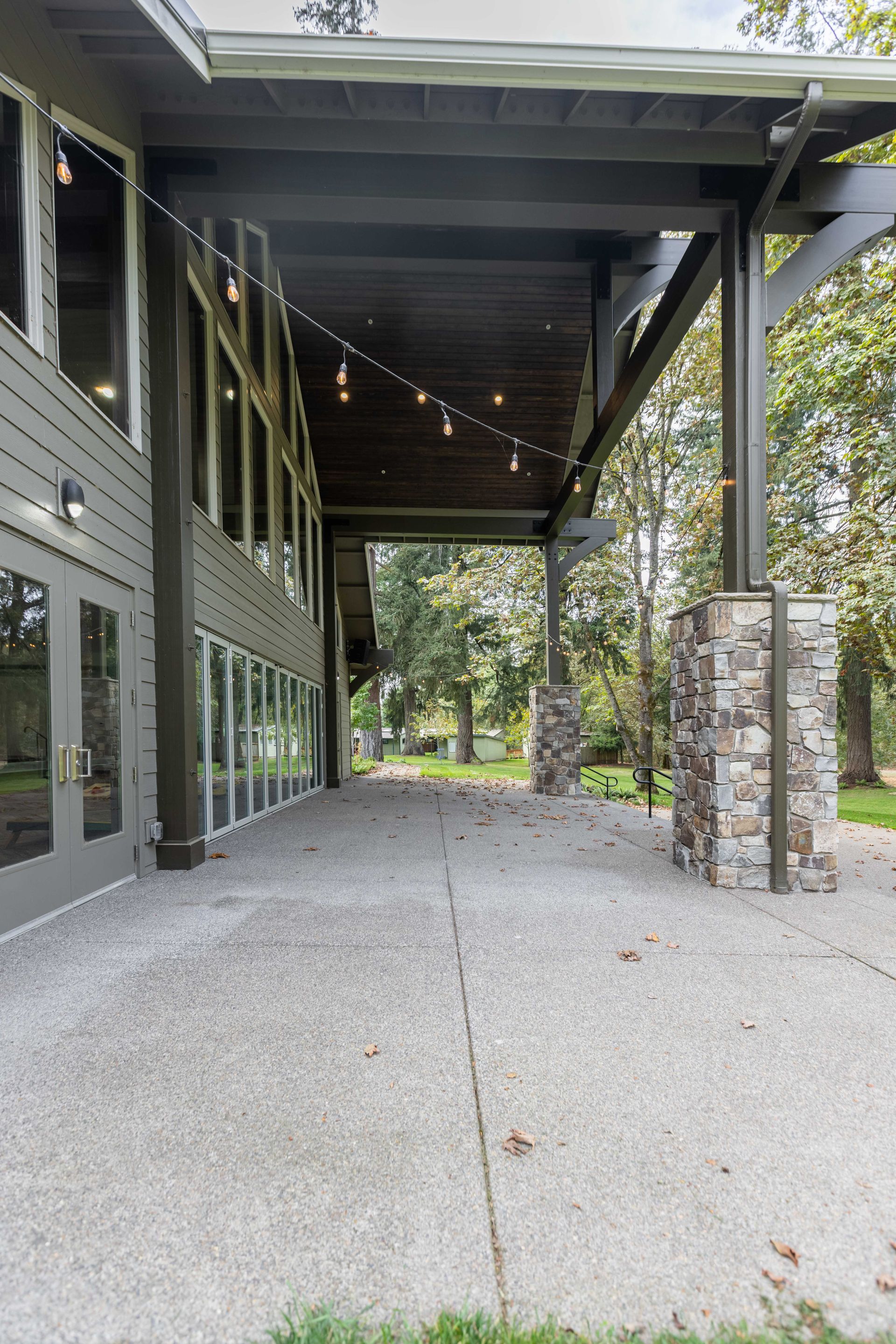 Covered outdoor patio with string lights, concrete floor, stone columns, and a building wall on the left