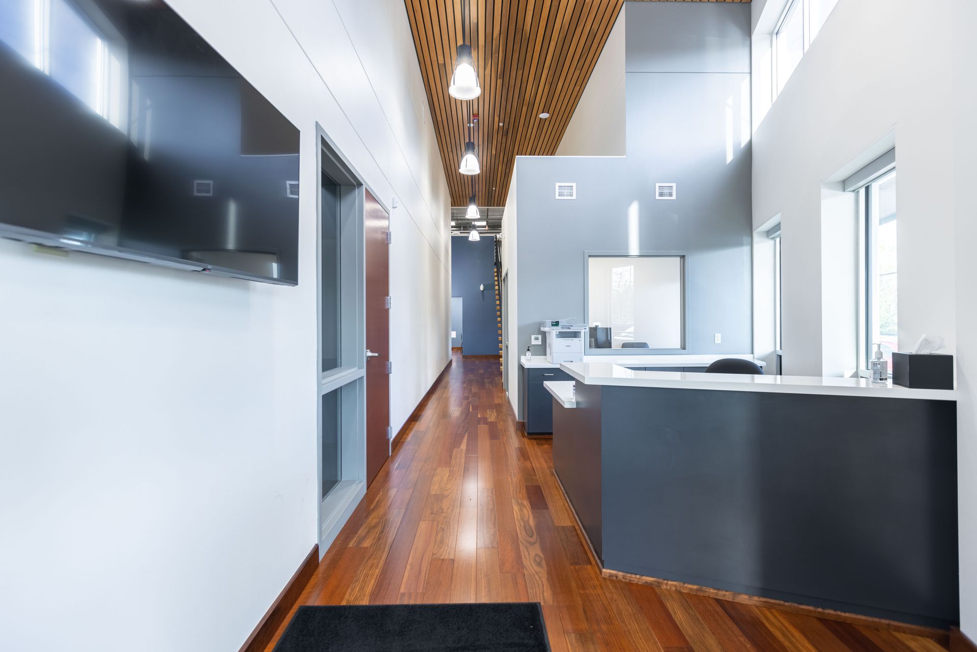 Modern office hallway with wooden floors, white walls, and dark reception desks under a wood-slatted ceiling
