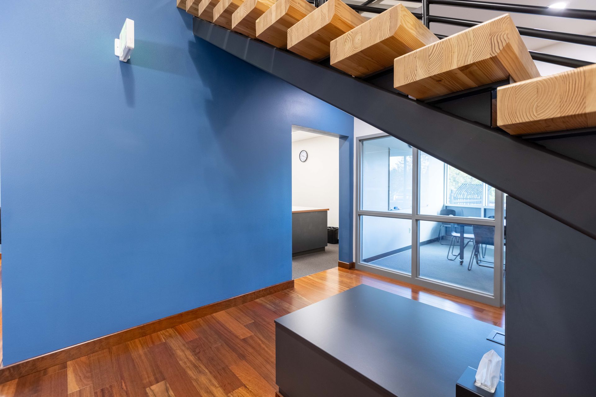 Modern office interior with blue wall, wood floor, glass doors, and a stairway overhead